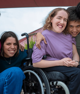 Three people smiling together outdoors. A woman in a wheelchair wearing a purple shirt has her arms around two friends on either side of her.