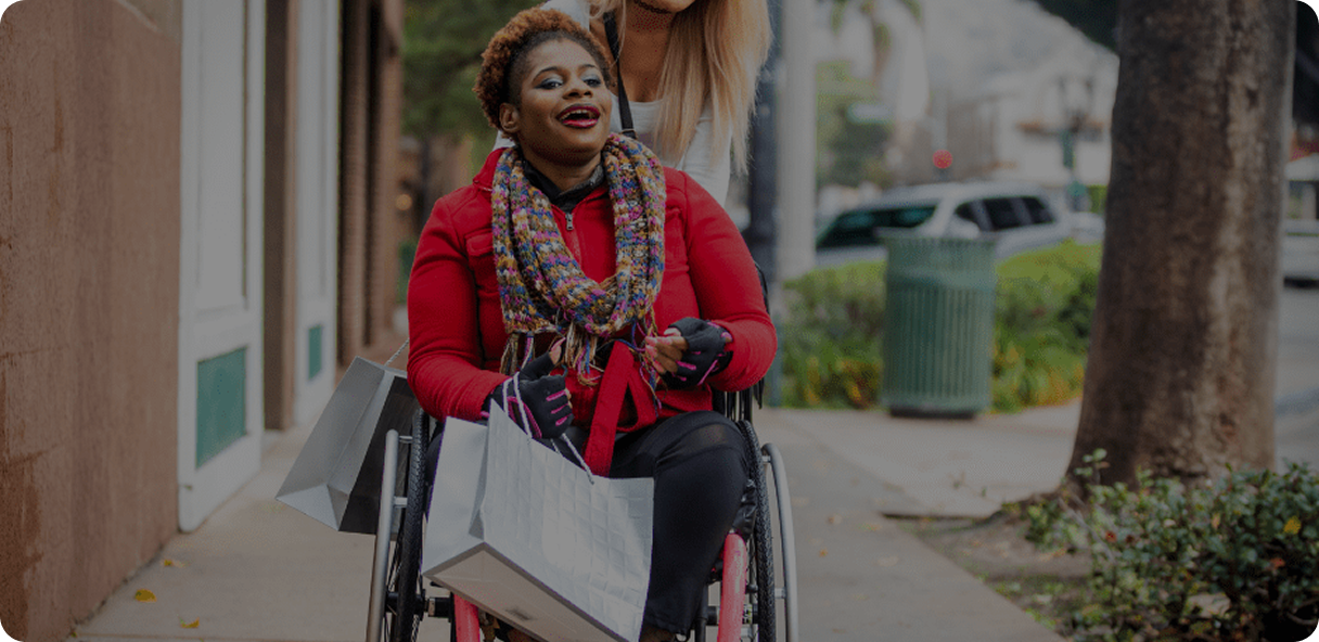 A smiling woman in a wheelchair holds shopping bags while being pushed by another person on a sidewalk lined with trees and buildings.