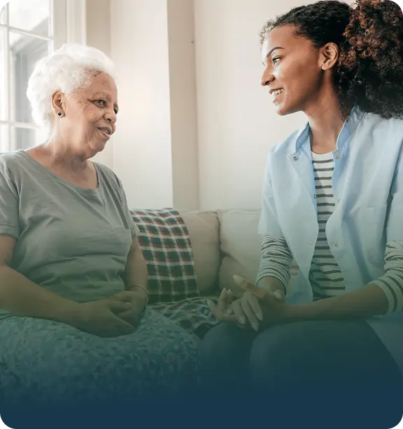 A young caregiver or nurse sits on a couch, smiling and talking with an older woman in a comfortable, sunlit room. Both appear engaged and relaxed.