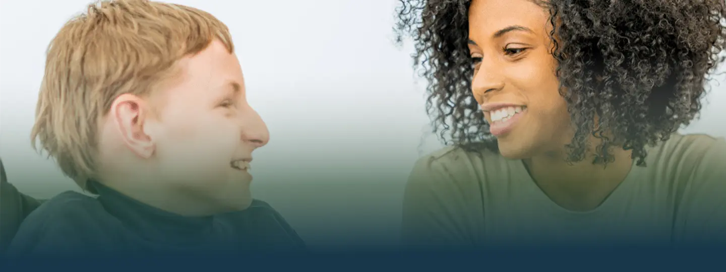 A young boy with light brown hair smiles at a woman with curly dark hair, who is also smiling at him. They appear to be having a positive and friendly conversation.