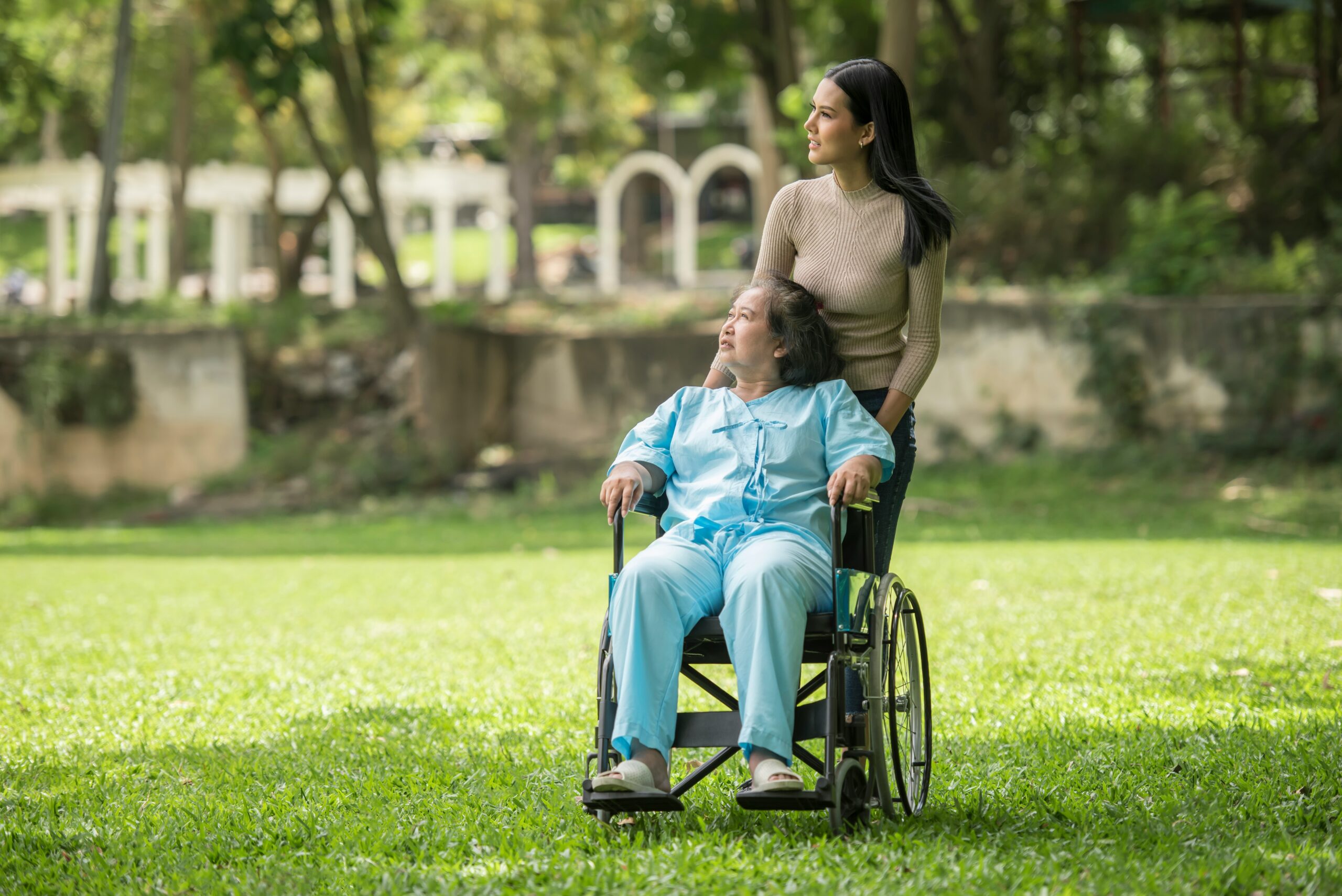 A young woman stands behind an older woman in a wheelchair. They are outdoors on grass, surrounded by trees, both looking to the side and appearing content, embodying the support and care often found through OPWDD provider resources NY.